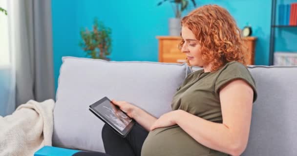 Mujer embarazada es relajante en la sala de estar en el sofá con la tableta. Ella ha recibido — Vídeos de Stock