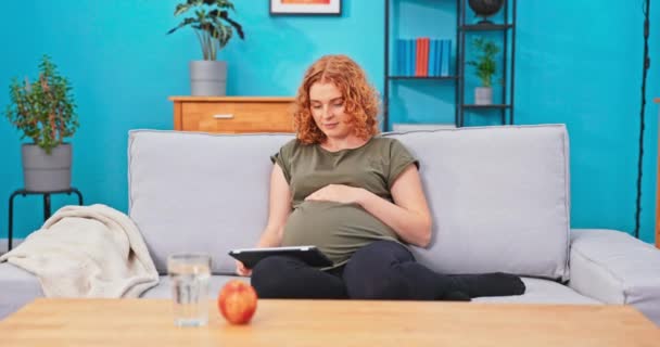 Una mujer embarazada pelirroja se relaja en el sofá de la sala de estar, sostiene la tableta en el regazo y navega — Vídeos de Stock
