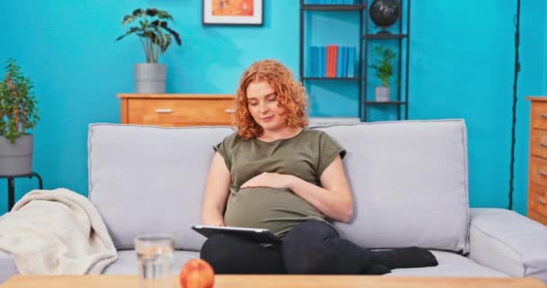 Красивая девушка с беременным животиком отдыхает на диване в гостиной — стоковое видео