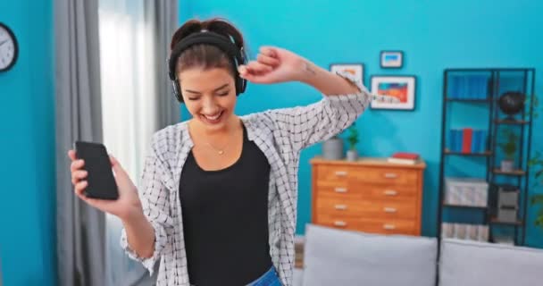 Uśmiechnięta kobieta tańcząca w pokoju, bezprzewodowe słuchawki na uszach, trzymająca telefon, machająca głową — Wideo stockowe