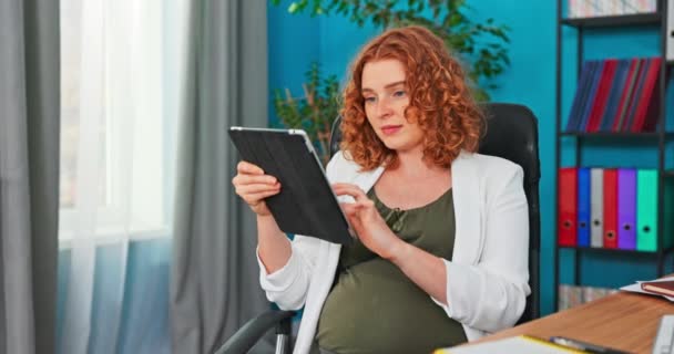 Uma jovem mãe senta-se no escritório no trabalho enquanto faz uma pausa segurando um tablet — Vídeo de Stock