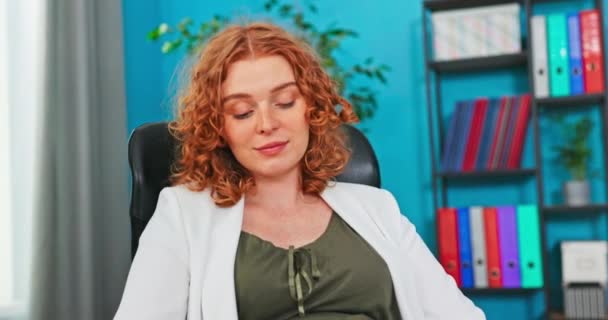 Hermosa mujer con el pelo rojo rizado está descansando en una silla en compañía, vestida con — Vídeo de stock