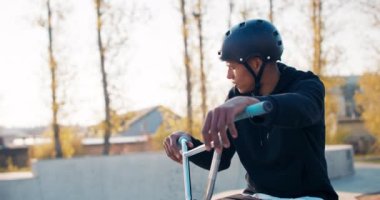 Gevşek kazak giyen ve siyah kasklı bir adam rampanın tepesinde BMX 'in üzerinde dinleniyor.,