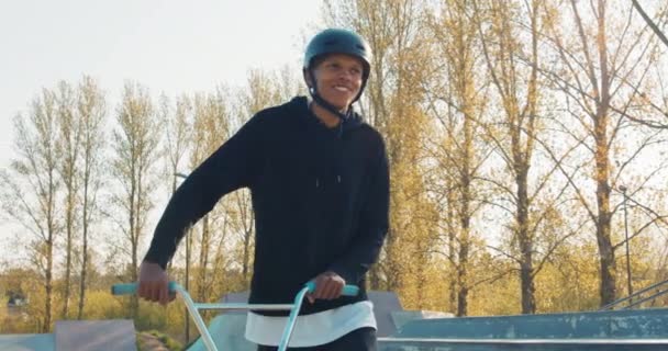 Entertaining student spending active time after college, walking through skatepark leading bike, helmet on head, — Stock Video