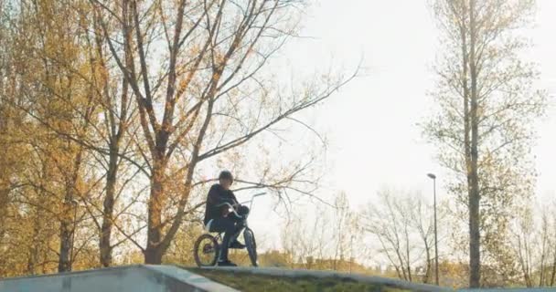 Um jovem estudante sorri enquanto ele se senta com bicicleta em uma rampa e um capacete — Vídeo de Stock