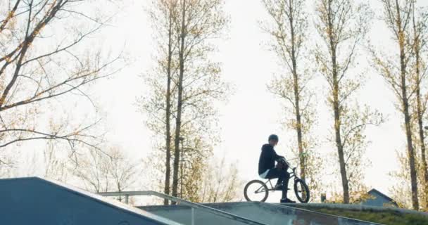 Um jovem estudante sorri enquanto ele se senta com bicicleta em uma rampa e um capacete — Vídeo de Stock