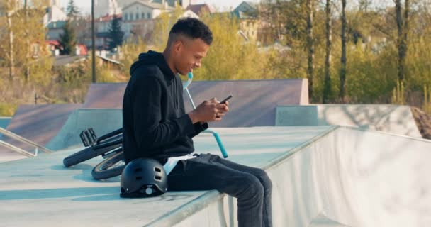Ciemnoskóry nastolatek siedzi na betonowej rampie, rower za nim, trzyma telefon w rękach, słupki na — Wideo stockowe