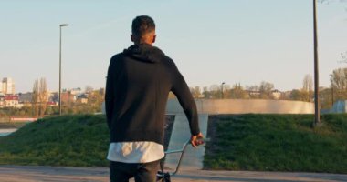 Sokak kıyafetleri giymiş esmer bir çocuk BMX 'i yanında tutuyor ve rampaya tırmanıyor.