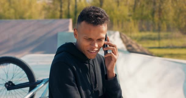 Ciemnoskóry przystojny nastolatek siedzi w skateparku po południu, bmx obok niego, telefon trzymany do — Wideo stockowe