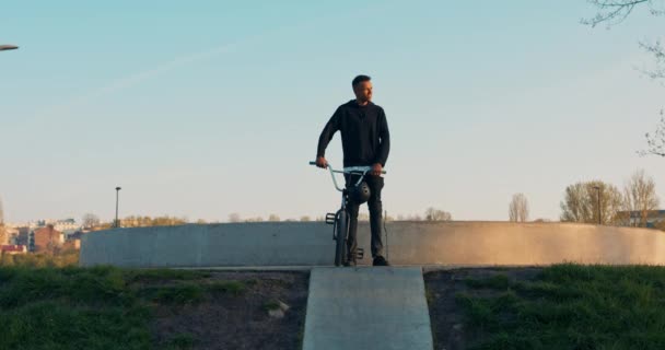 Joven ciclista principiante, de pie en la rampa, sosteniendo la bicicleta y el casco en la mano, vestido todo en — Vídeos de Stock