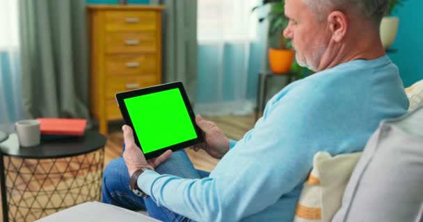 Ein erwachsener alter Mann zu Hause benutzt ein grünes Mock-up-Tablet. Er sitzt auf einem — Stockvideo