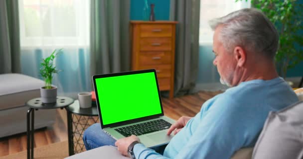 Ældre mand derhjemme Sidder på en sofa Arbejder på en bærbar computer med grøn – Stock-video