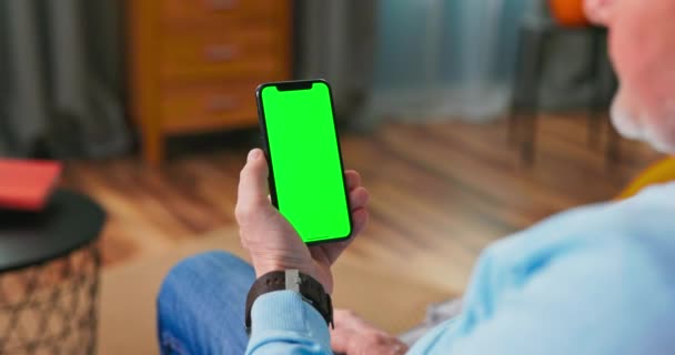Man thuis Zittend op een bank met Smartphone met Green Mock-up Screen. Kerel gebruikt — Stockvideo