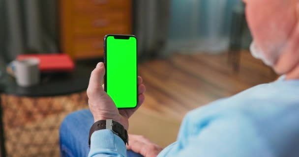 Homem mais velho em casa sentado em um sofá funciona em um telefone celular com verde — Vídeo de Stock