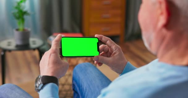 Äldre man hemma vilar på en soffa Använda med grön Mock-up Screen Smart Phone — Stockvideo