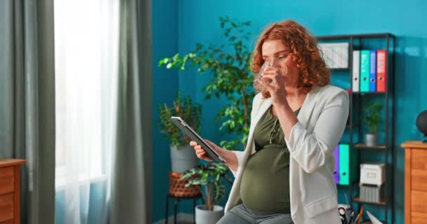 Frauen mit Schwangerschaftsbauch sitzen mit Tablette in der Hand am Schreibtisch, trinken Wasser aus Glas, — Stockvideo