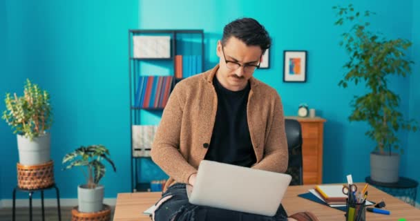Комфортно одетый мужчина, работающий в офисе сидит за столом с ноутбуком на коленях, амбициозный — стоковое видео