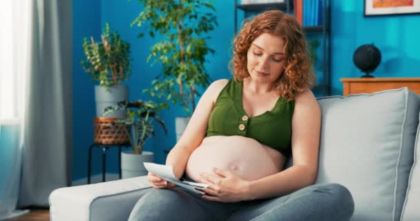 Mujer embarazada se sienta en el sofá en la sala de estar y sostiene su vientre, en la mano es — Vídeos de Stock