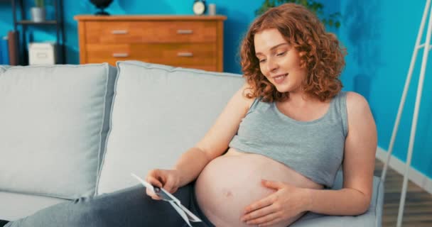Mujer satisfecha, vestida con ropa cómoda para el hogar, se encuentra en el sofá apoyado contra el reposabrazos, acariciando embarazada — Vídeos de Stock