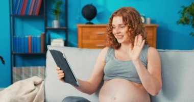 Kızıl saçlı kız elinde tabletle oturma odasındaki kanepede dinleniyor, doğum izninde, internette video sohbeti yapıyor, ailesiyle, arkadaşlarıyla kamerayla konuşuyor, selamlaşırken el sallıyor.