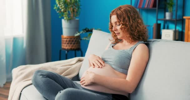 Femme attentionnée s'assoit sur le canapé avec les jambes croisées et parle au ventre enceinte, le caresse, — Video