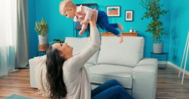 Genç, eğlenceli anne halıya oturur, bebeği kanepeden alır ve kaldırır.