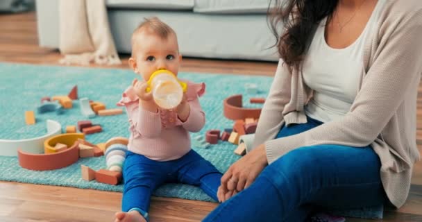 Sweet girl is sitting on carpet among toys with mother, holding bottle, cup in hands, — Stock Video