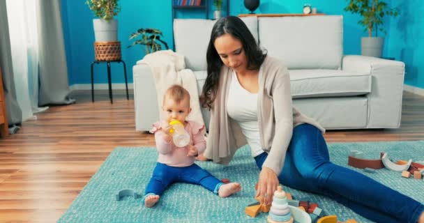 Mooi, klein meisje zit op tapijt tussen speelgoed met moeder, in handen houden fles, — Stockvideo