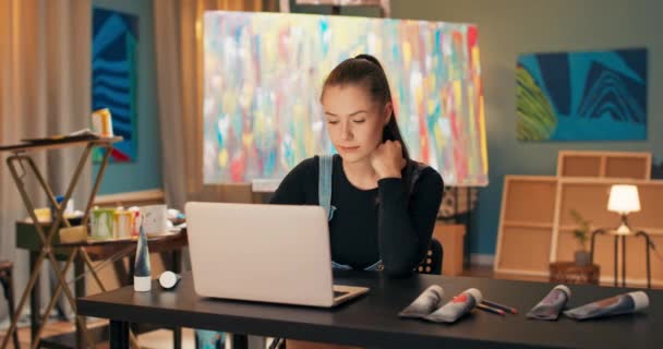 Uma morena sorridente senta-se em uma mesa na frente de um laptop no estúdio de arte — Vídeo de Stock
