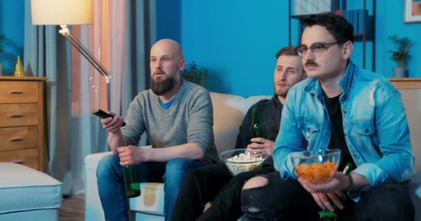 Tres mejores amigos pasan la noche juntos frente a la pantalla de televisión bebiendo cerveza comiendo papas fritas — Vídeos de Stock