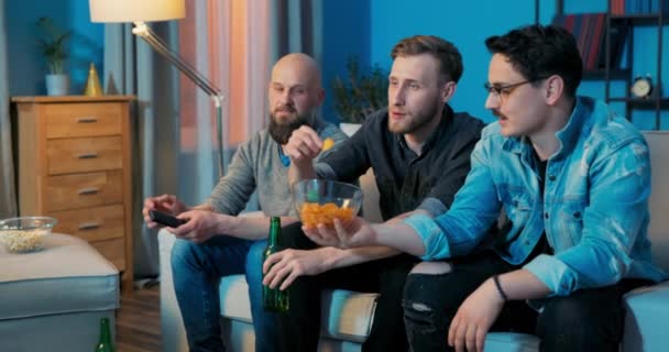 Mens kväll framför TV på soffan, killar dricker öl, äter popcorn chips pratar — Stockvideo