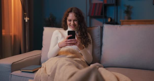 Una chica se sienta en el sofá por la noche a la luz de la lámpara, las piernas en el café — Vídeos de Stock