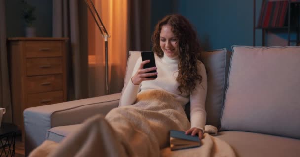 Una chica se sienta en el sofá por la noche a la luz de la lámpara, las piernas en el café — Vídeos de Stock