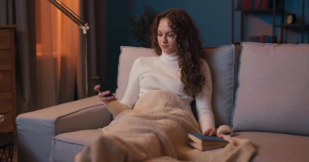 Vacker flicka sitter på soffan i ett bekvämt läge under filten tillbringar — Stockvideo