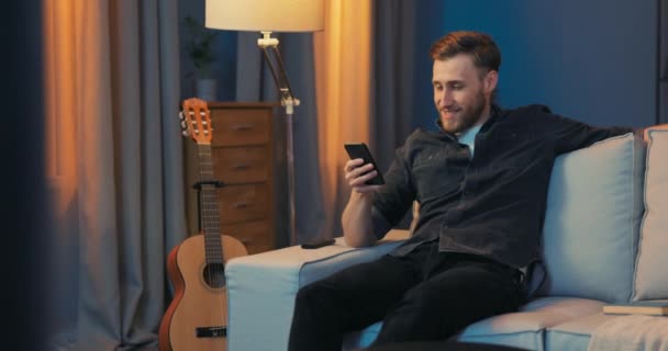 Молодой человек сидит довольный на диване в гостиной по вечерам — стоковое видео