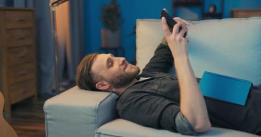 Sıkılmış bir adam oturma odasında kanepede uzanıyor, kafası duvara yaslanmış.