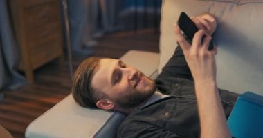 Sıkılmış bir adam oturma odasında kanepede uzanıyor, kafası duvara yaslanmış.