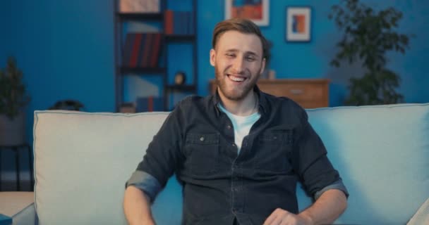 Расслабленный улыбающийся мужчина сидит на диване в гостиной, одетый в равнину — стоковое видео
