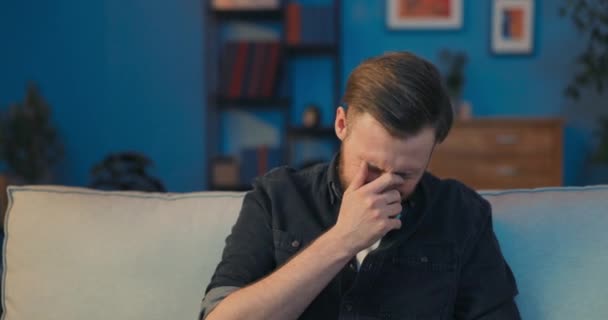 Relajado hombre sonriente se sienta en el sofá en la sala de estar, vestido en una llanura — Vídeos de Stock