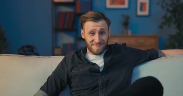 Un hombre guapo está sentado en el sofá tarde en la noche frente a la pantalla de TV — Vídeos de Stock