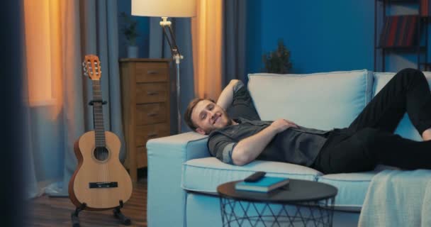 An attractive man lies comfortably on the sofa in front of TV, relaxing after a — Stock Video