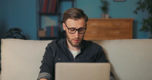 Молодий співробітник, IT-спеціаліст, одягнений в окуляри, сидить у вітальні увечері — стокове відео