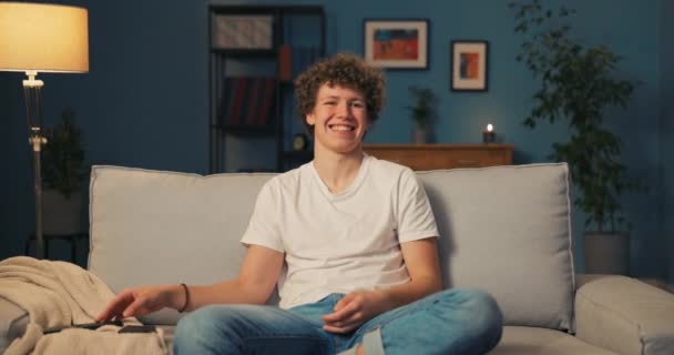 Un chico morena sonriente con el pelo rizado se relaja en el sofá de la sala de estar en el — Vídeos de Stock