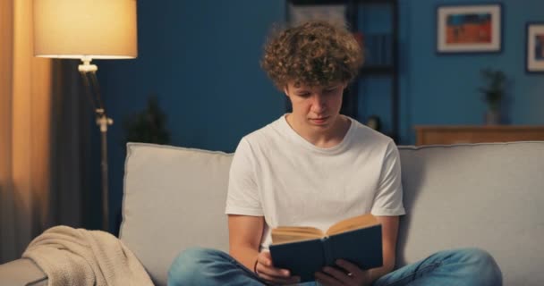 Hombre guapo con pelo afro se relaja en traje hogareño mientras profundiza en el libro que es — Vídeos de Stock