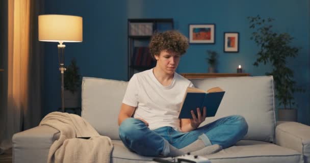 Um jovem adolescente relaxa no sofá lendo um livro escolar, o livro aborrece — Vídeo de Stock
