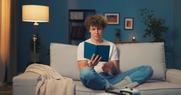 Um jovem adolescente relaxa no sofá lendo um livro escolar, o livro aborrece — Vídeo de Stock