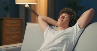 Genç bir adam kanepede dinleniyor, elleri başının arkasında, televizyonu kapatıyor ve dinliyor.