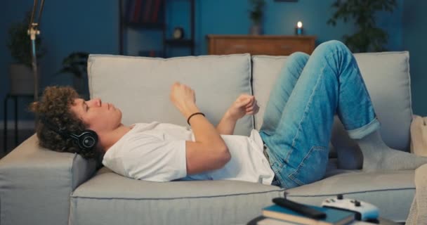 Mladý chlapec s kudrnatými vlasy leží doma na gauči a poslouchá — Stock video