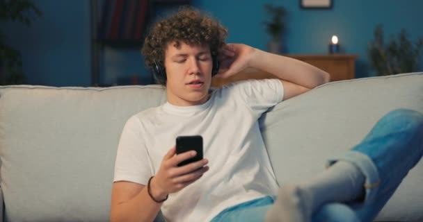 Hombre atractivo con el pelo rizado se sienta en un sofá con la pierna metida, auriculares inalámbricos — Vídeos de Stock