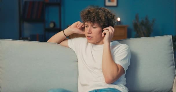 Un adolescente con el pelo rizado se sienta en un sofá de la sala de estar con auriculares inalámbricos — Vídeos de Stock